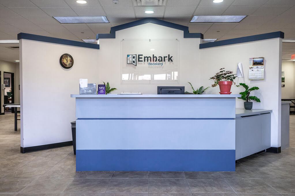 Front Desk with Embark Logo sign on wall, clock, plants and monitor